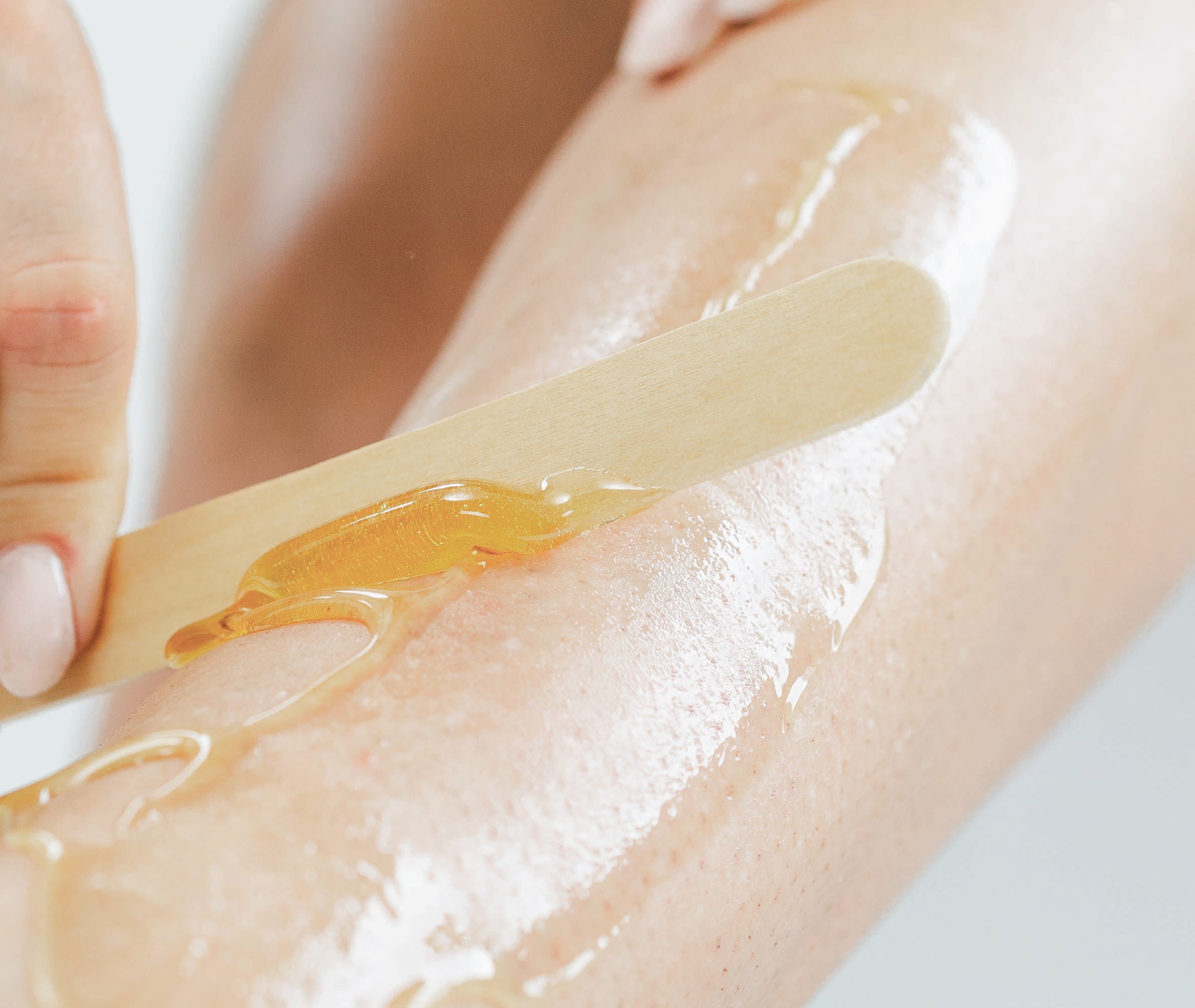 close up of person applying Kiss Waxing strip sugar to their leg with a wooden spatula on a light background